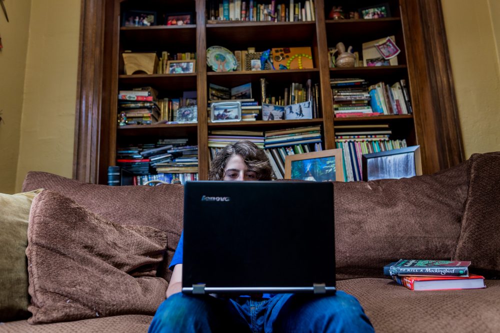 Gabe Fleisher is sitting on a brown couch and looking at a black Lenovo laptop opened in front of him