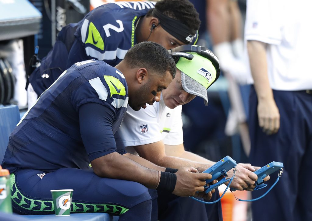 Russell Wilson and a coach sit on the sidelines of a preseason game looking at a Surface tablet.