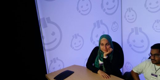 Woman in a hijab sitting at a table with a man, looking up at a screen