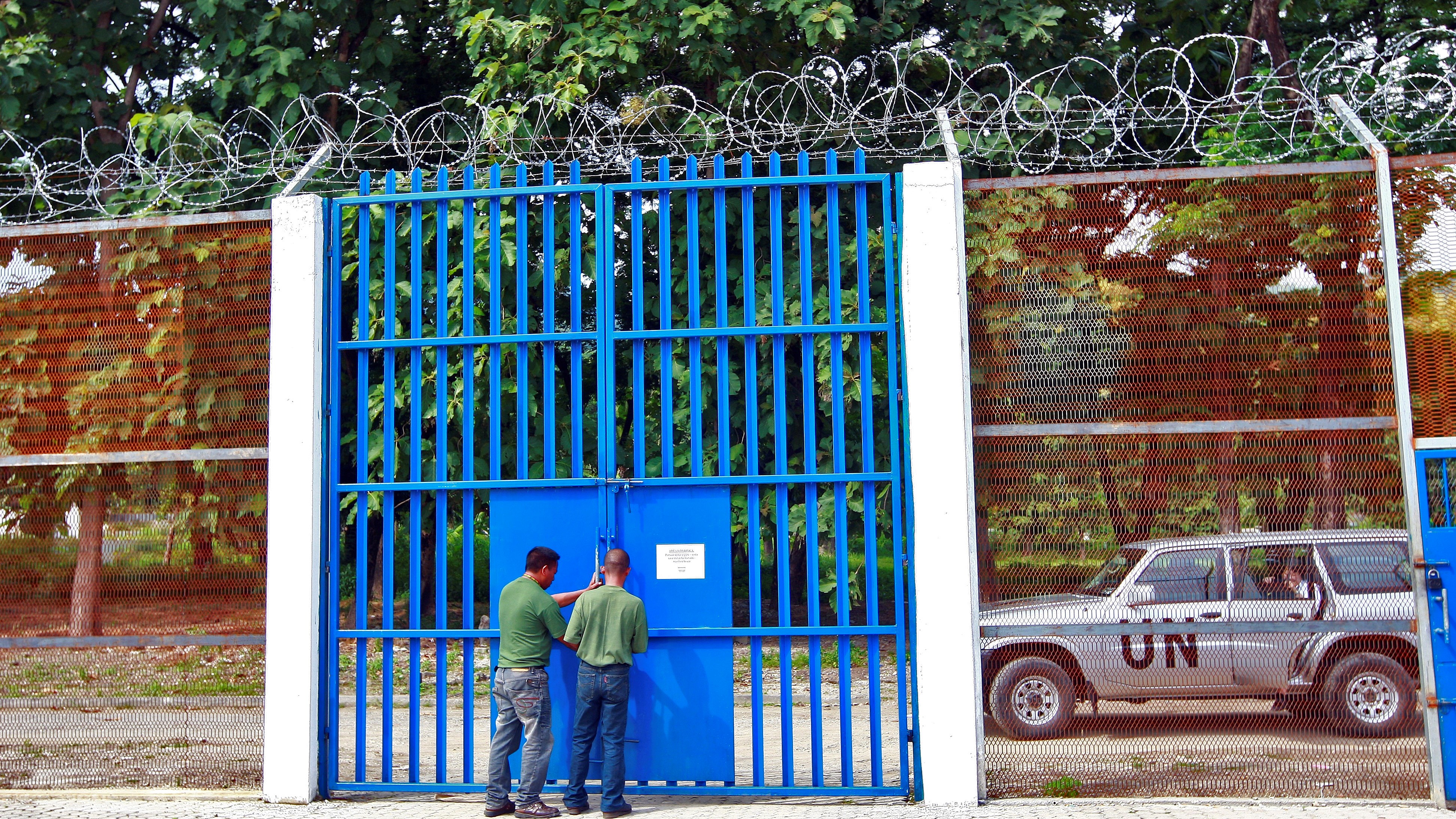 Photo of two workers standing at a security gate with a UN vehicle in the background