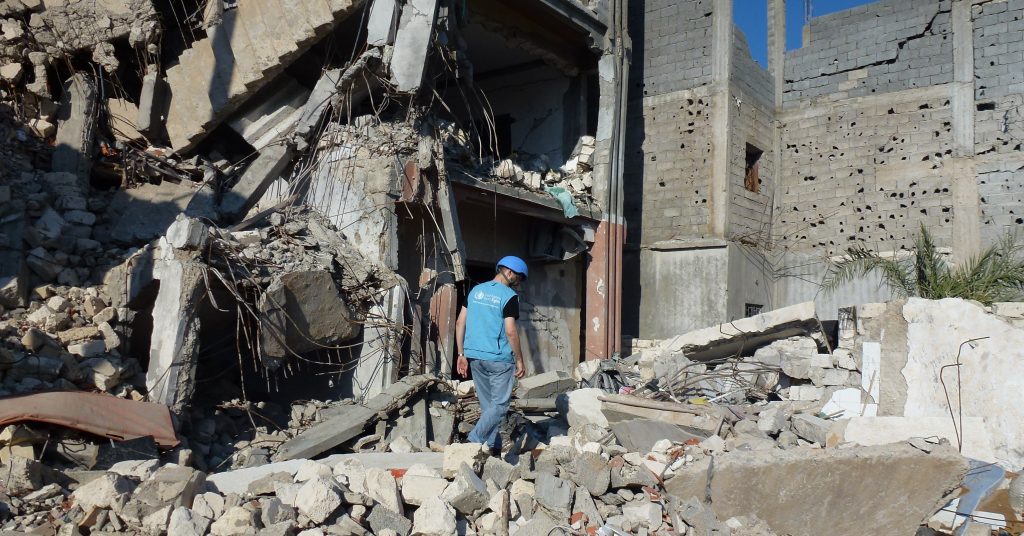 Photo of man in vest and hard hat walking over chunks of concrete near a destroyed structure