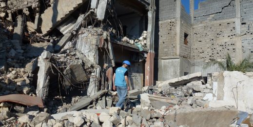 Photo of man in vest and hard hat walking over chunks of concrete near a destroyed structure