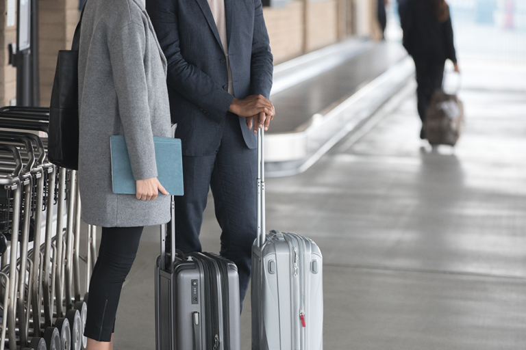 Two people stand with their suitcases at an airport curb