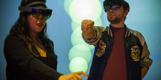 A woman and a man, both wearing HoloLens sets, make pinching motions in the air to place virtual "blooms" while bubbles float on the screens around them