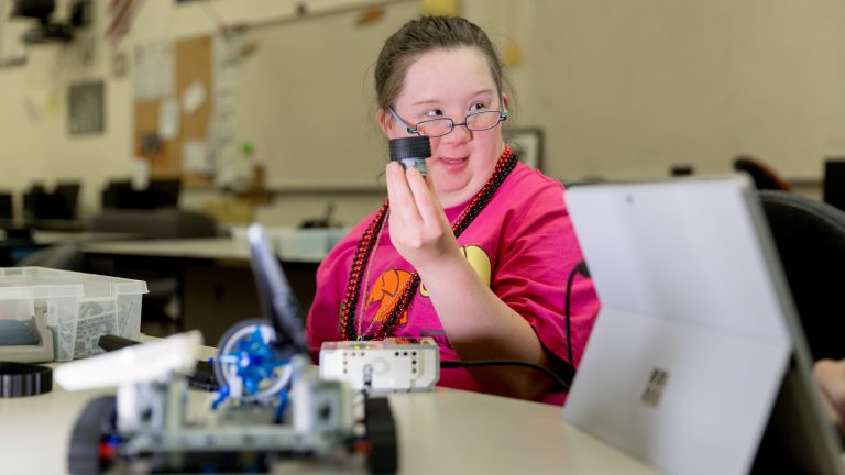 Photo of girl sitting in front of a Legos robot and holding up a wheel.