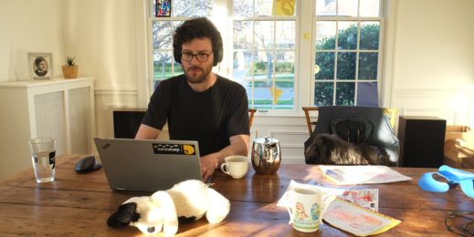 man sits in front of laptop at table with dog, toys and coffee cup around him