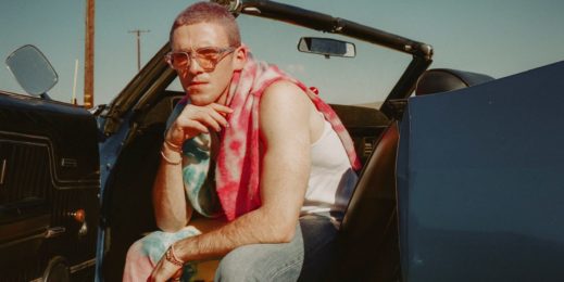 Lauv wears a pink tie-died scarf, pink sunglasses, and dyed pink hair. He leans out the driver's side of a classic convertible.