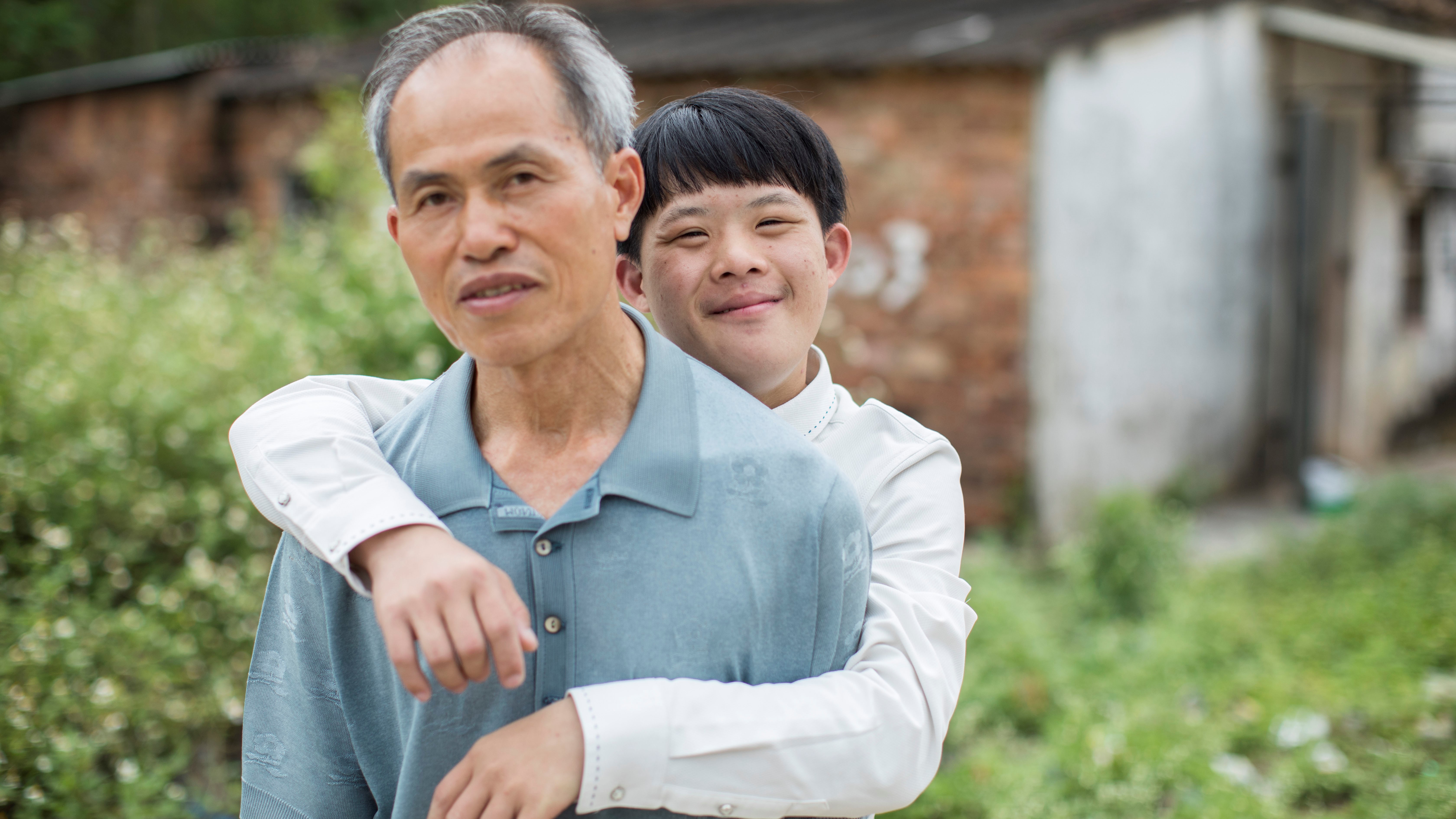 Photo of boy standing behind father with arms around him.