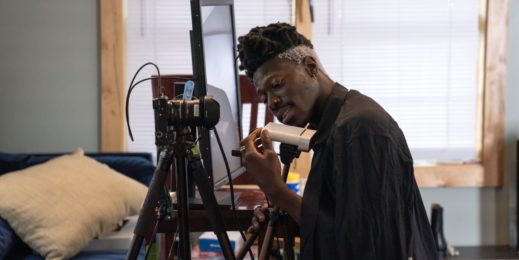 Musician Moses Sumney kneels to examine the Azure Kinect hardware, whi ch is on a tripod next to a mounted camera and a table with a large computer monitor. A blue sofa is in the background; we are in his living room