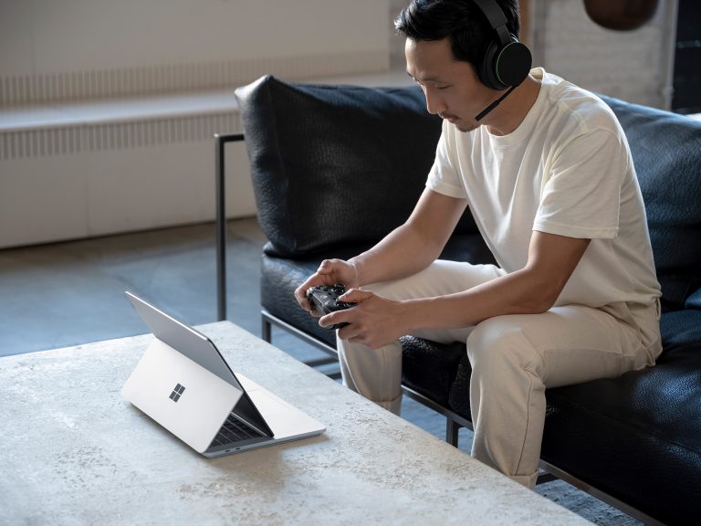 A man sitting down using an Xbox controller with a headset on while on a Surface laptop