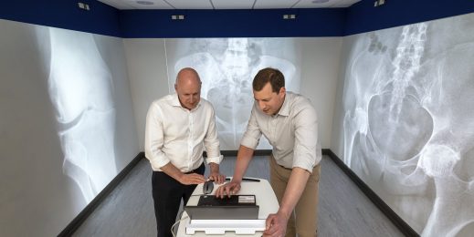 Two men dressed in smart trousers and shirts stand in a small room. They are standing in front of a laptop and are looking at the screen. Large images of knee and hip joint X-rays have been projected onto the walls behind them and either side of them. 