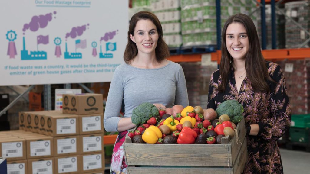 Two women standing behind a wooden box filled with fresh vegetables