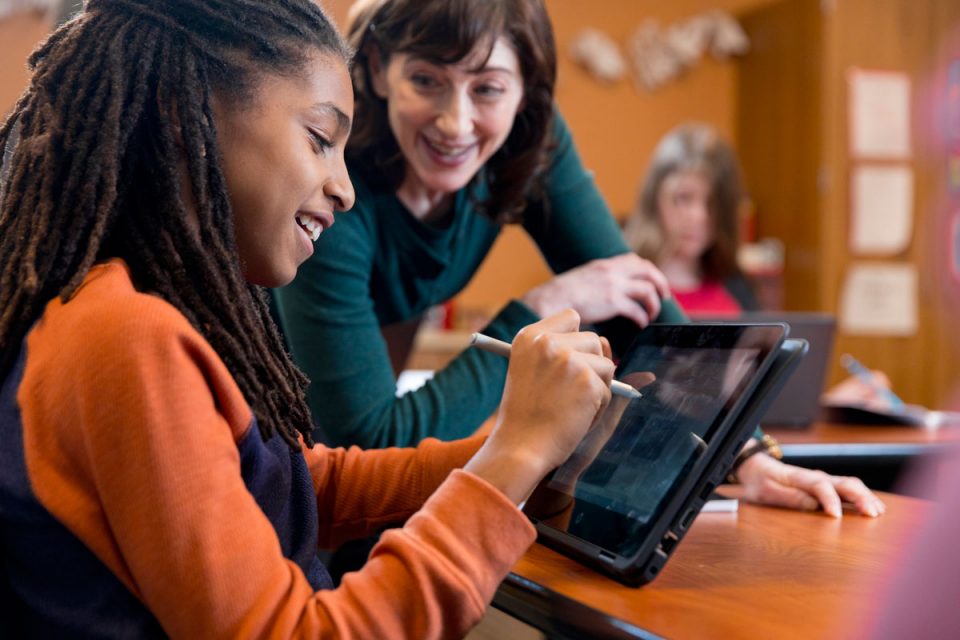 Female teacher speaks with female middle school student who is sitting at desk in classroom, using pen on screen of convertible laptop folded open as tablet.