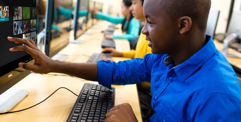 Education Color Photo of male student touching device screen with keyboard on desk and students in background at computer lab
