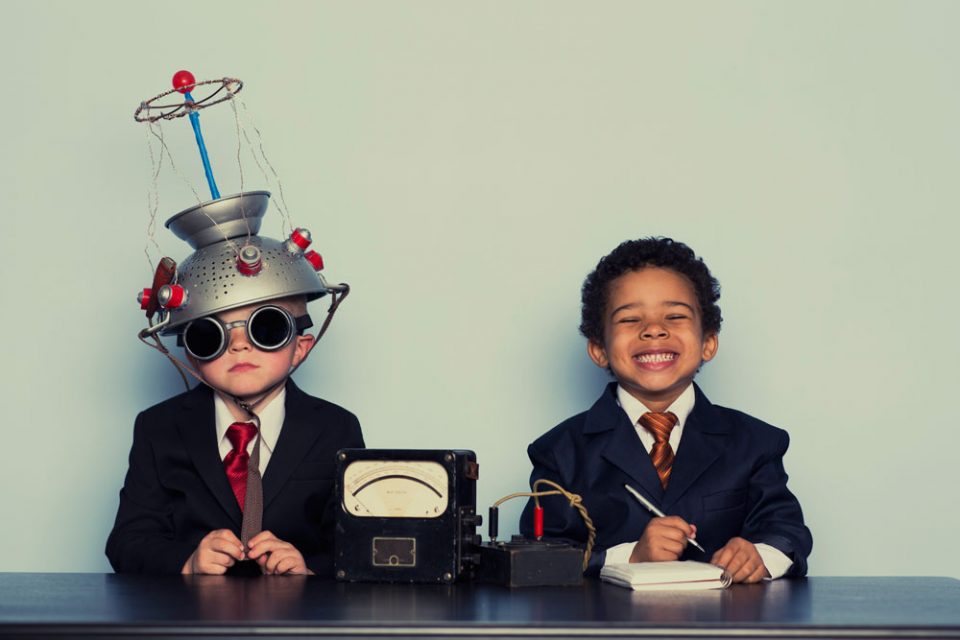 Two boys sitting behind a table laughing. Boy on the left has a technical device on his head. Education. Hour of Code campaign.