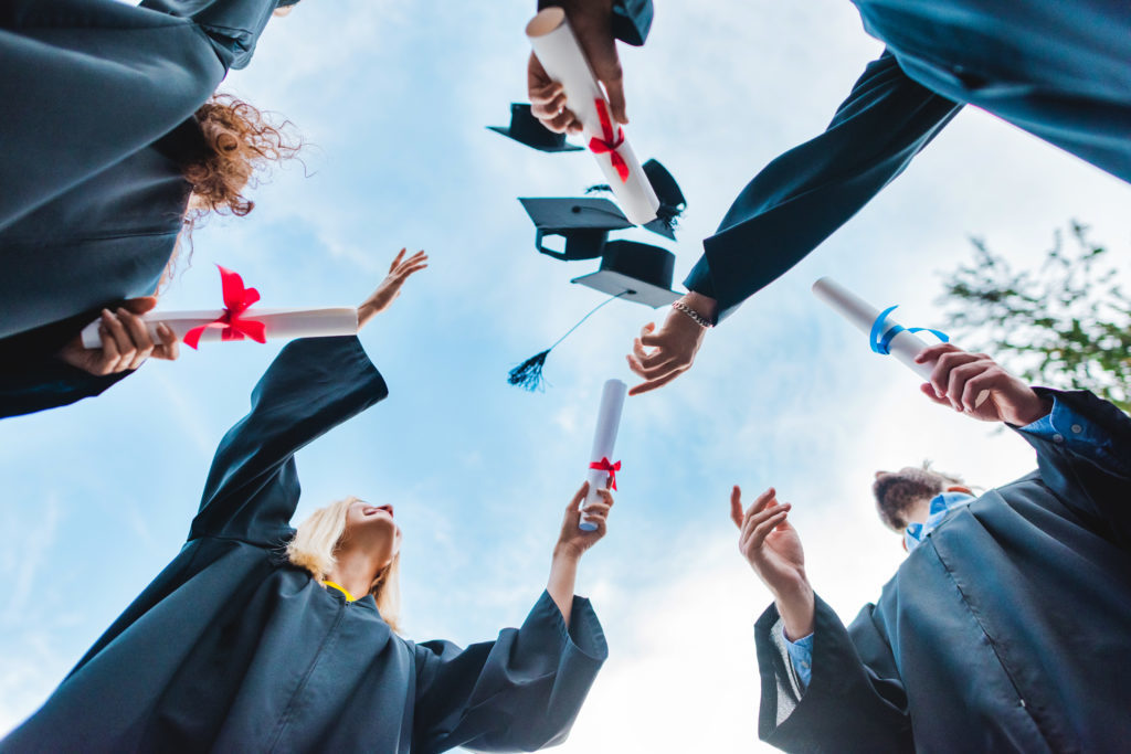 bottom view of happy multicultural graduates with diplomas throwing caps up with blue sky on background