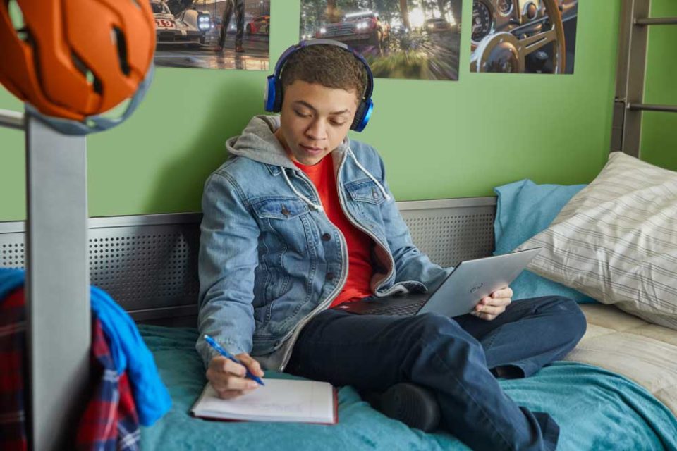 Boy sitting on bed with a laptop on his lap writing in his workbook