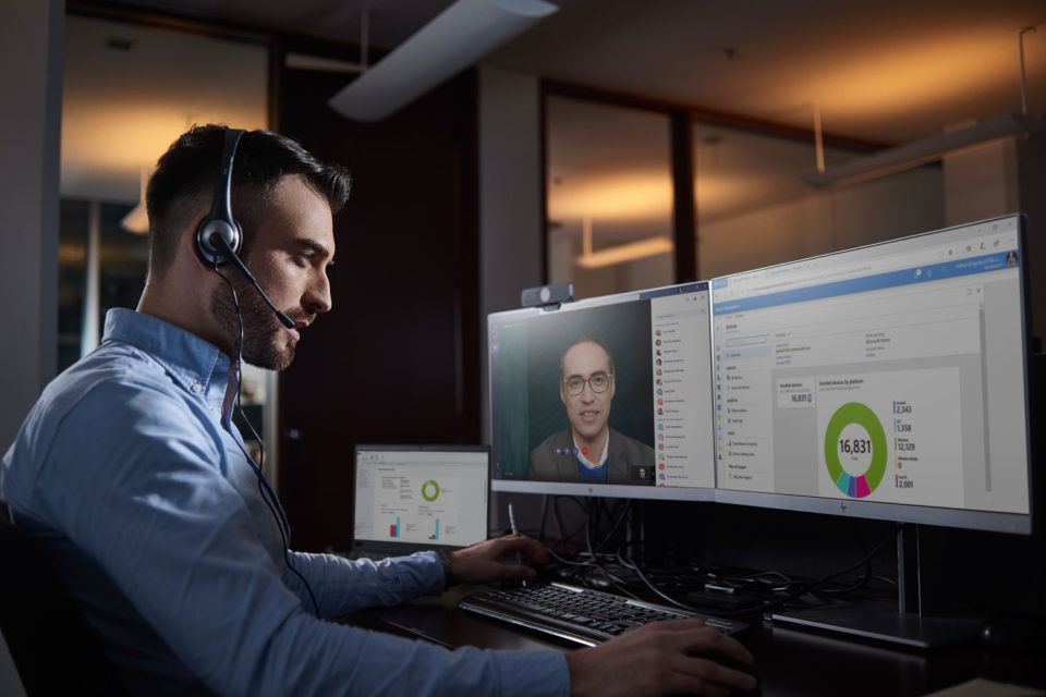 Man sitting at his desk with headphones on talking to another man on a computer monitor