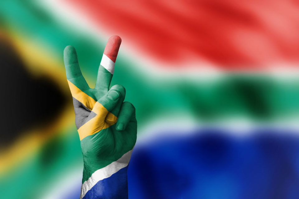 South African flag in the colours red, blue, black, green, gold and white in the background and painted on a person’s hand who is holding a peace sign.