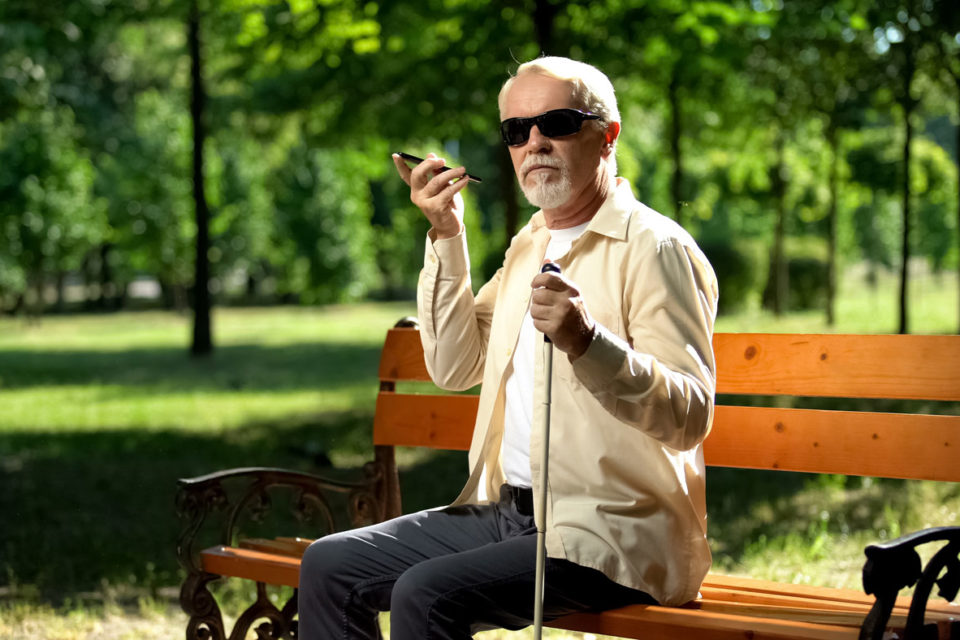 Man with visual impairment sits on park bench listening to mobile narration.