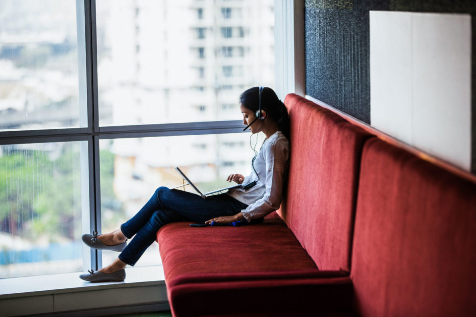 A woman with headphones on working at her laptop while sitting on a red couch next to a window in a sky rise building.