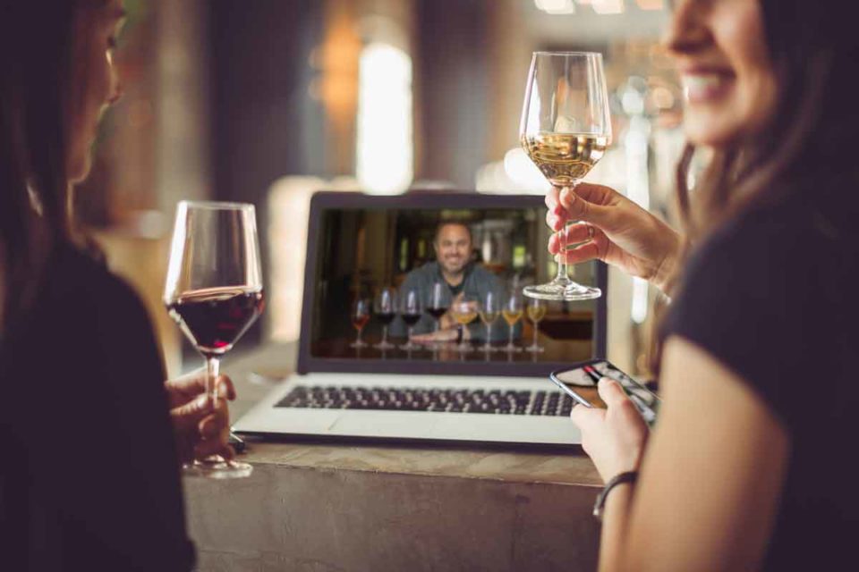 Two ladies, smiling, holding a wine glass, with a laptop open. On the Laptop screen a man is smiling while eight wine glasses are in front of him.