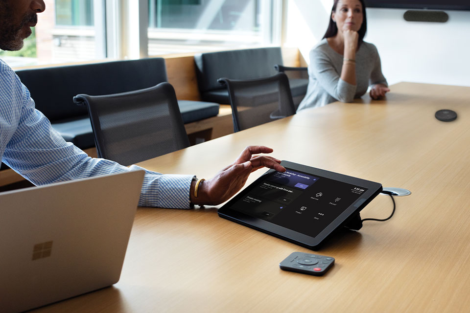 One male and one female in a large conference room with the male engaging a Teams Meeting Rooms touch display preparing to join a Teams Meeting