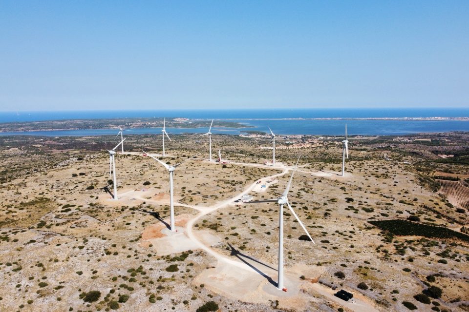 Eight wind turbines top an arid hill, with the Mediterranean Sea in the distance.