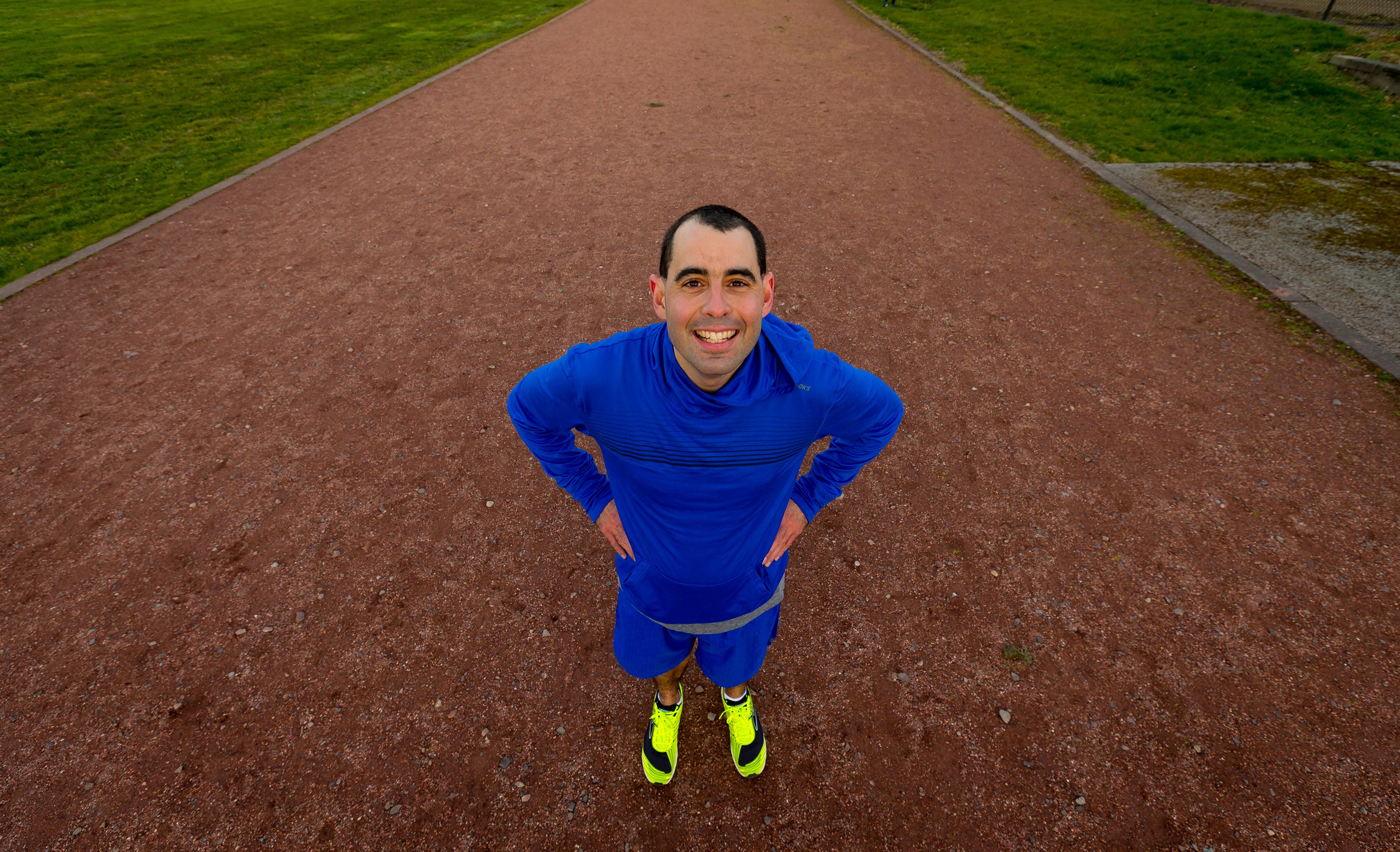 photo of man in a bright blue running outfit and yellow shoes grinning with hands on his hips in the middle of a red running track