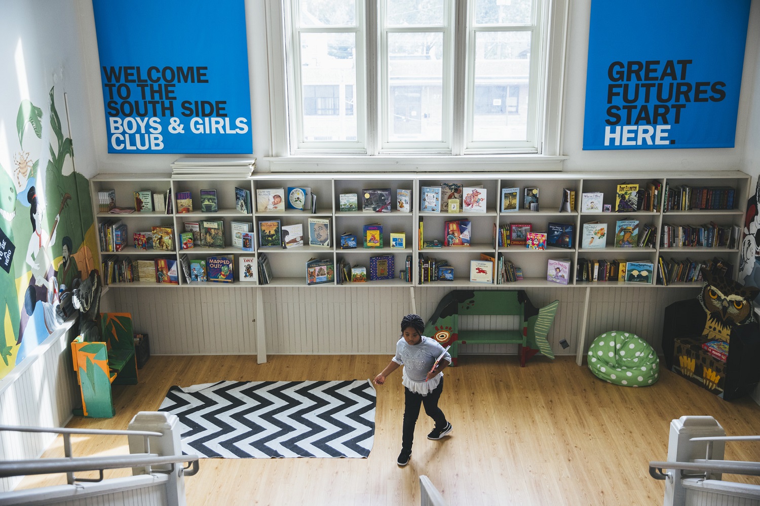 A young girl walks near a staircase in a building with high ceilings and big windows