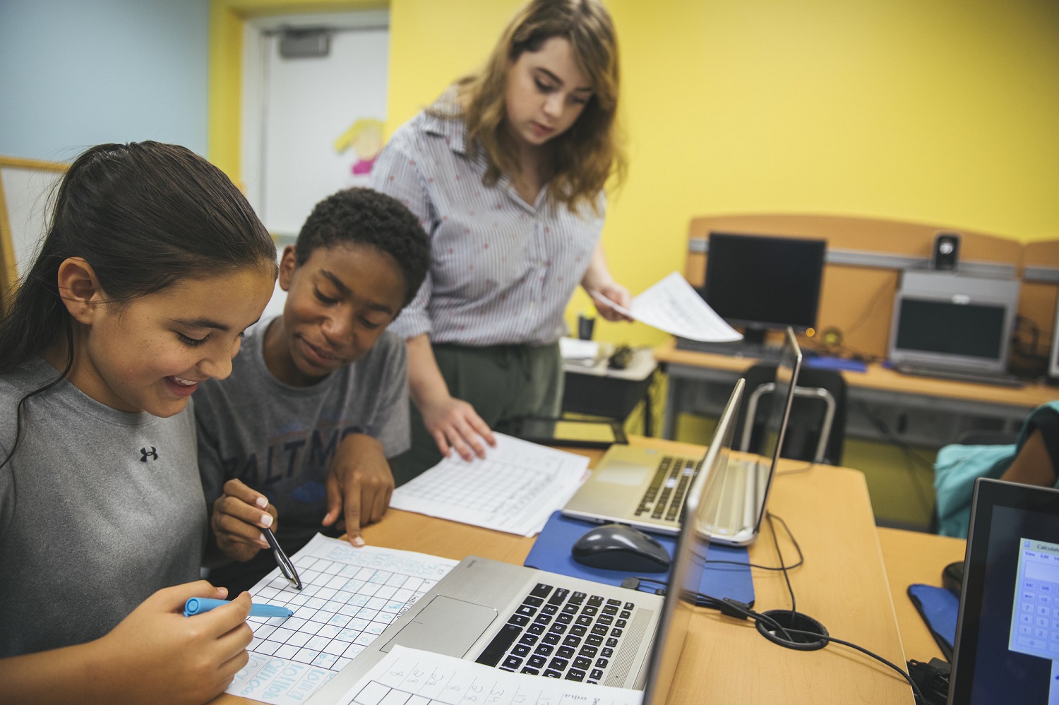 A girl and boy smile while working in a computer room with a female teacher standing nearby.
