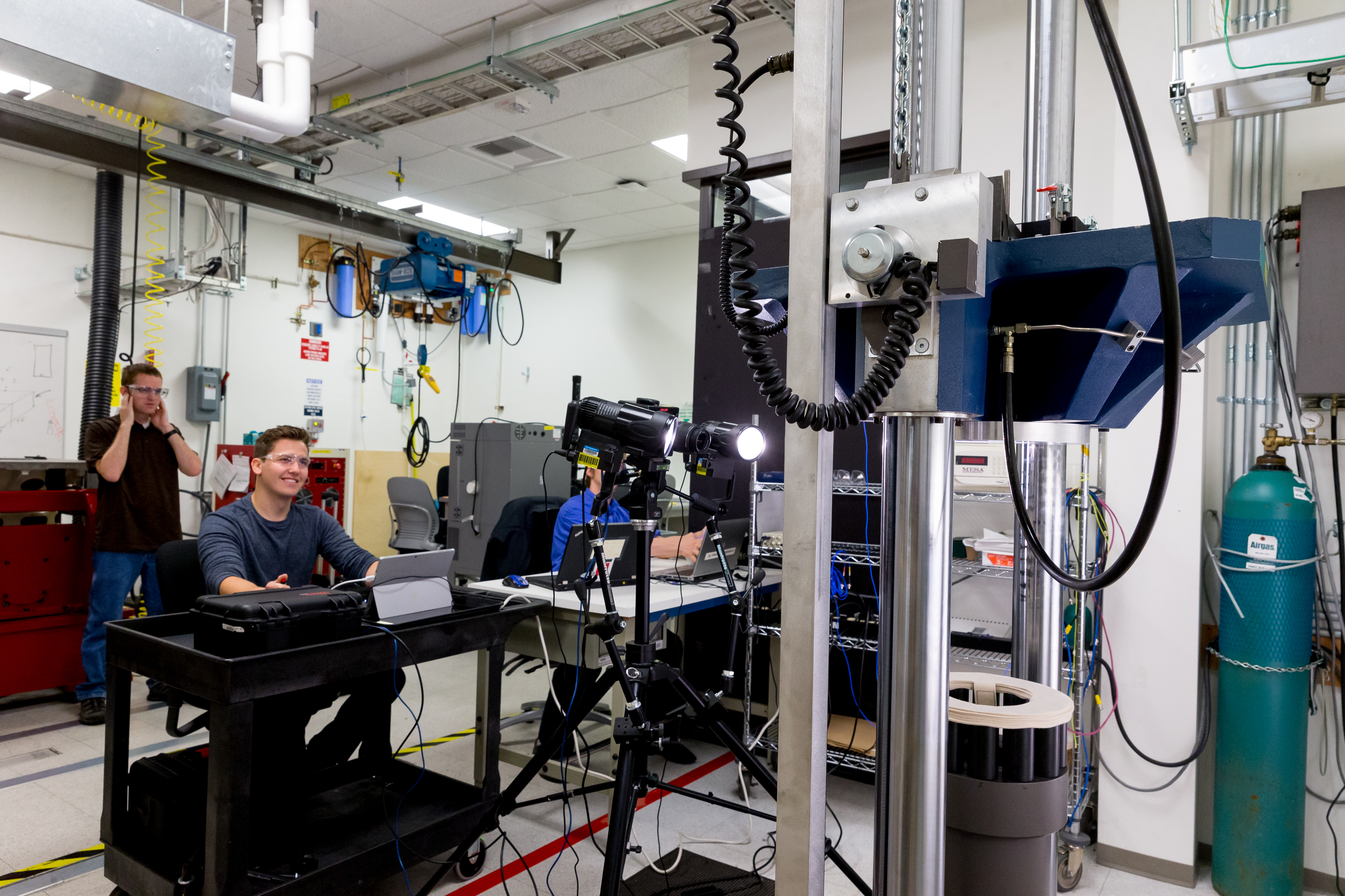 Technicians wearing ear and eye protection conduct an impact test on a packaged Surface device, simulating a 48-inch drop onto wood. Reliability test engineer Michael Nist stands behind reliability technician Vladimir Artym. Reliability engineer Jacob Murphy sits partially obscured by a test camera.