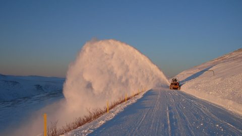 Machine blows arc of snow off a snowy road against blue sky