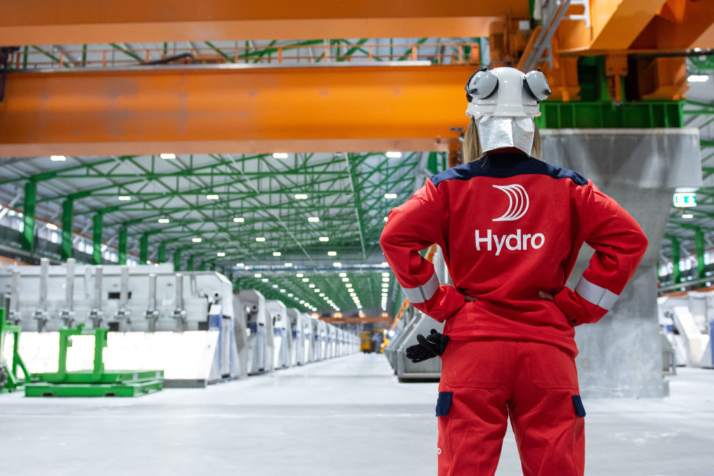 A worker is facing away from the camera and wearing a red Norsk Hydro jacket while looking out at a factory floor.