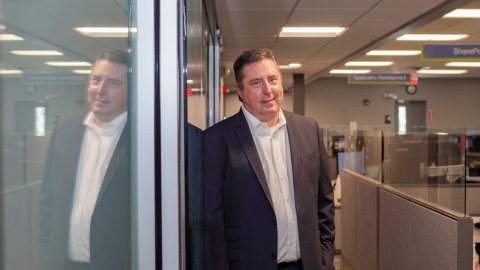 A man in a dark sport jacket and white shirt leans against a glass door, which does his reflection.