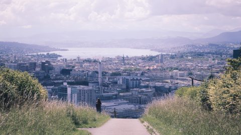 The city of Zurich sits below a bank of clouds. In the fore ground, a road is flanked by green grass.