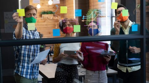 A team of co-workers diagram a problem on a clear white board.