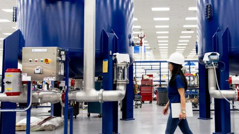 Female worker walking along factory floor in manufacturing plant, in front of two large blue vats.