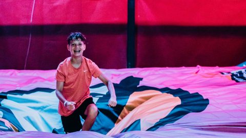 A boy stands, smiling, in the middle of a large soft cushion after falling from a climbing attraction at an indoor amusement park.