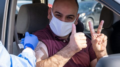A man wearing a mask and sitting in a car gets a COVID-19 vaccination from a health care worker. The man is holding up two fingers on his left hand to signify receiving his second dose. He is also sporting a thumbs up sign with his right hand.