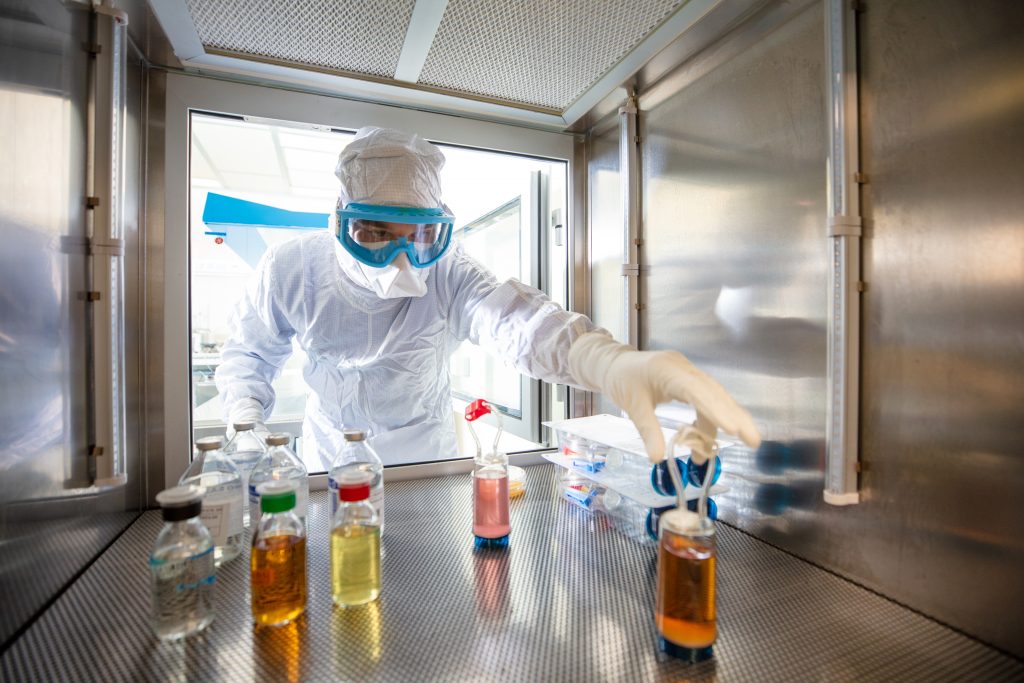 A scientist wearing a white protective suit and head covering reaches a gloved hand into an air-tight hood containing bottles of medicine.