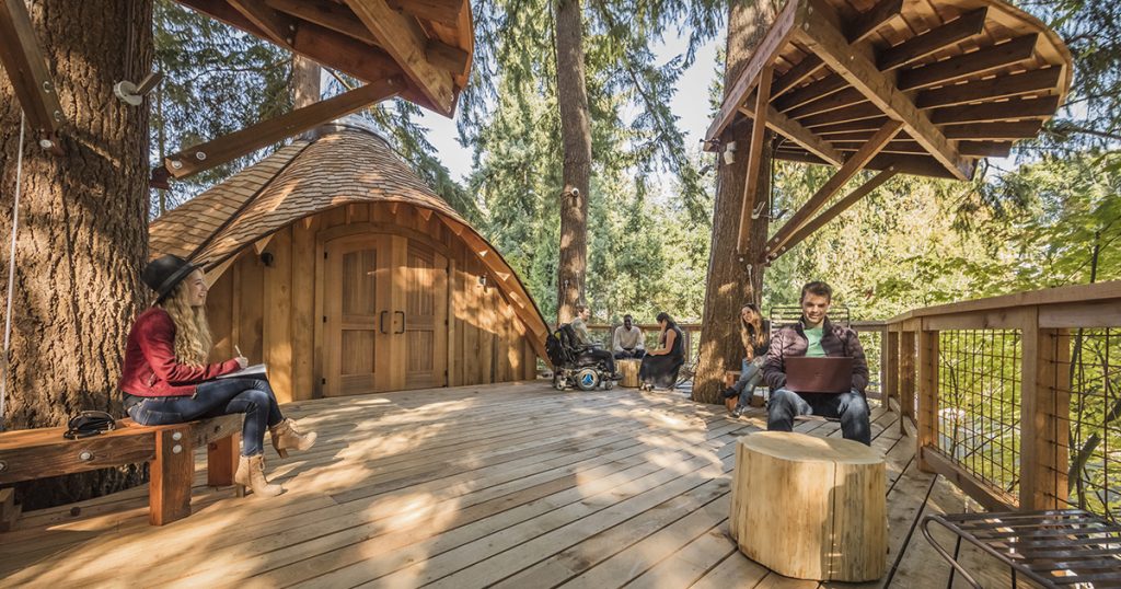 Employees with computers and notebooks sitting on chairs and benches on a wooden deck surrounded by trees