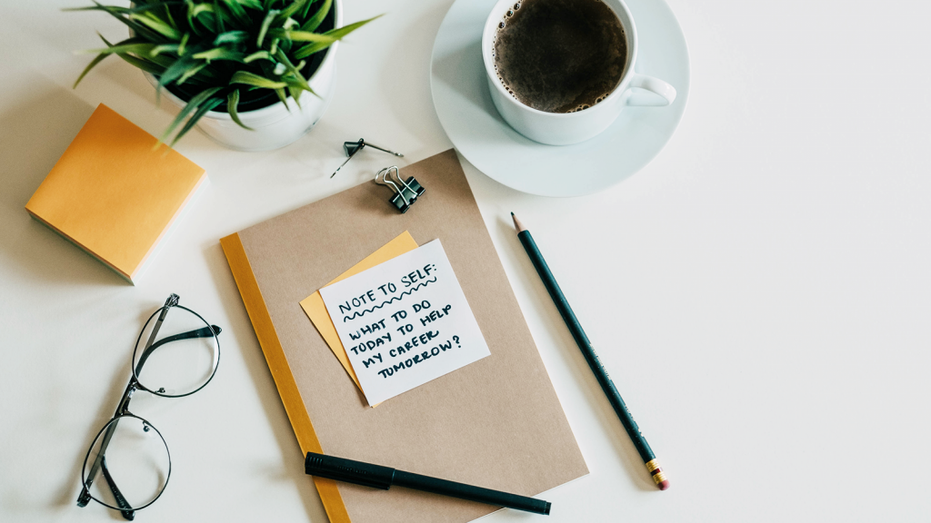 A desk with a cup of coffee, a plant, and a notebook with the words 'note to self: what to do today to help my career tomorrow' on it