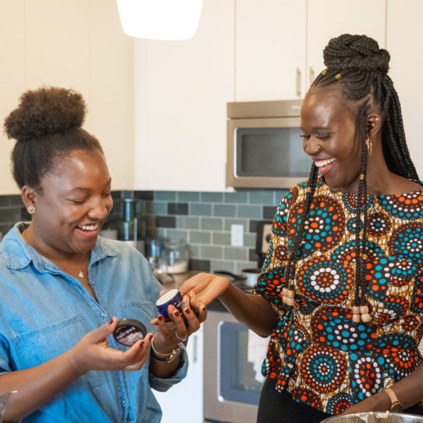 Two women in a kitchen laughing as they hold a jar of shea butter