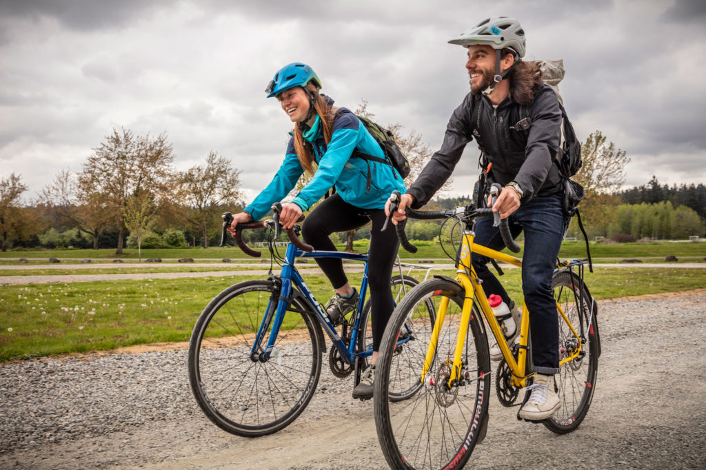 A man and a woman each ride a bike on a gravel trail