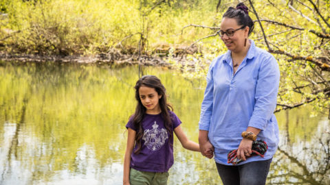 A mother and daughter hold hands by the river and smile