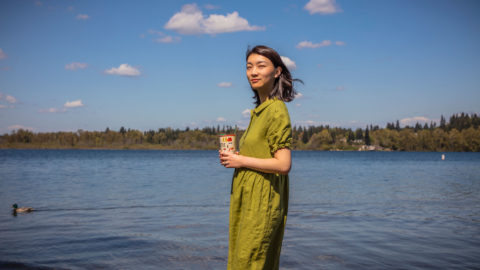 A woman in a green dress who is holding a coffee cup and standing at the shore of a lake