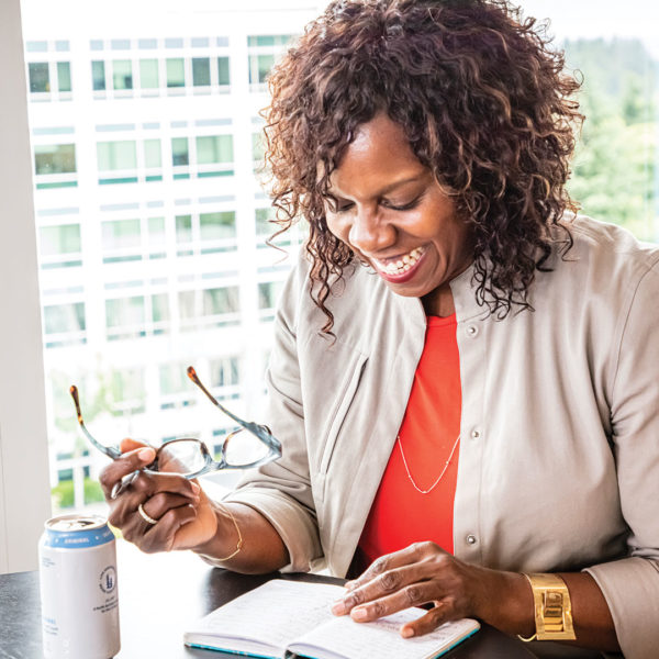 A smiling woman sitting at a desk holding reading glasses and looking at a notebook