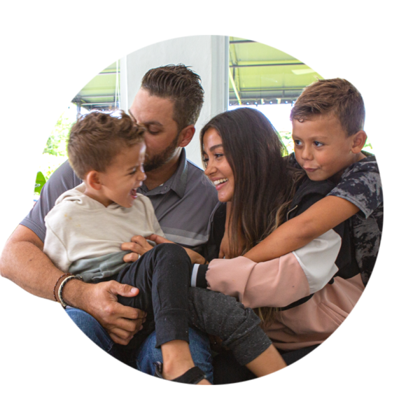 A woman tickles a little boy sitting in her husband's lap while her other son hangs on her sholders