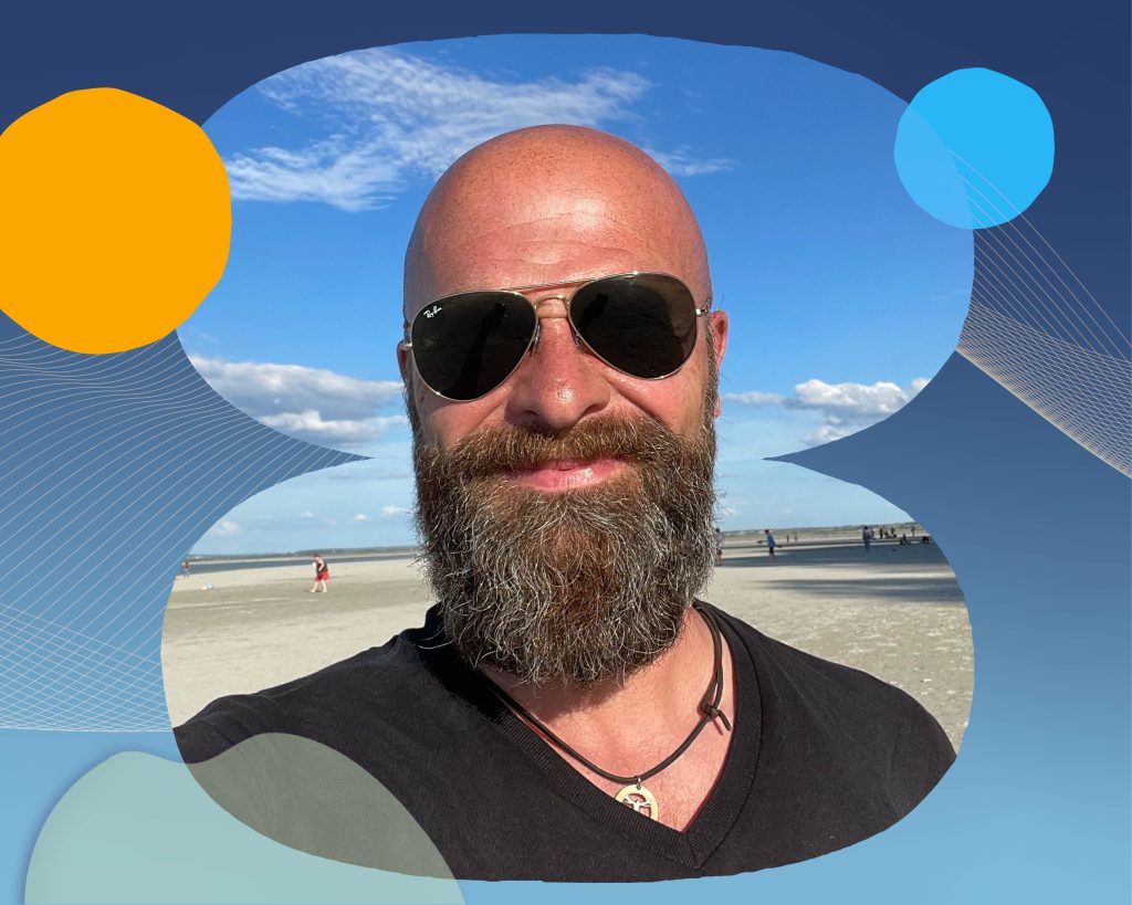 Sascha outside wearing sunglasses. He has a long beard and is smiling at the camera.